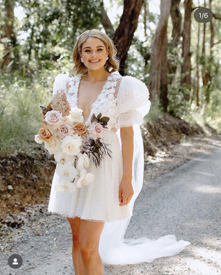 Ethereal white bridal dress with intricate floral appliqués, dramatic puff sleeves, and a playful short hemline, designed for relaxed outdoor weddings or unique bridal styles