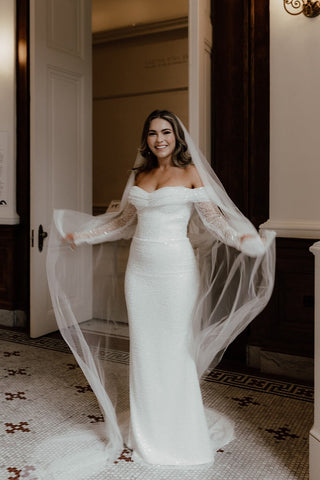 Elegant off-the-shoulder bridal gown with a fitted sequin design, sheer lace long sleeves, and a floor-length silhouette, paired with a flowing veil, perfect for glamorous wedding celebrations