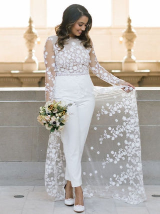 Elegant bridal jumpsuit featuring a sheer lace top with floral embroidery, long sleeves, and fitted white trousers, complemented by a flowing floral lace overskirt, perfect for modern weddings