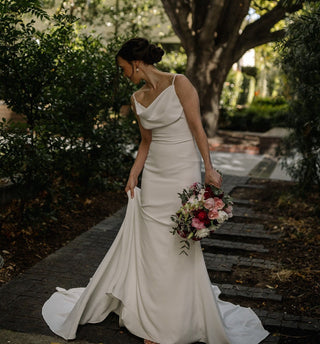 Elegant white wedding dress with a timeless cowl neckline, draped bodice, and a sleek, fitted silhouette that flows into a delicate train