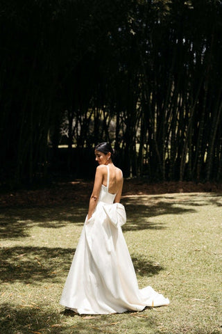 Chic outdoor wedding look with a classic white gown, thin straps, flowing train, and bold oversized bow at the back, captured in a lush garden setting