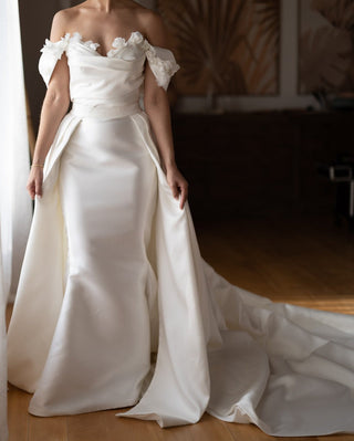 Ivory satin off-the-shoulder wedding gown with floral applique detailing, a fitted silhouette, and a dramatic floor-length train, perfect for elegant and timeless bridal looks