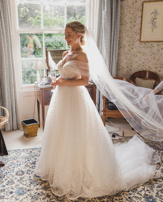 This gown features a romantic off-the-shoulder design with delicate draped tulle, creating an ethereal and timeless look