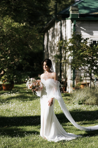 Graceful off-the-shoulder wedding gown featuring sheer tulle sleeves, a fitted silhouette, and a chapel train, perfect for a romantic outdoor ceremony