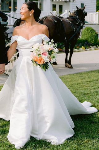 Timeless off-the-shoulder bridal gown with a cinched waist and dramatic pleated skirt, offering a regal and sophisticated silhouette