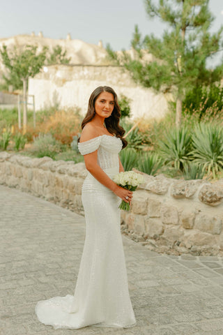 Elegant off-the-shoulder wedding dress featuring a beautifully beaded fabric that hugs the body, ideal for a chic and sophisticated bridal style.