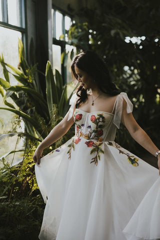 Ethereal white dress featuring vibrant floral embroidery across the bodice and skirt, complemented by delicate tulle straps for a romantic, garden-inspired look