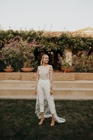 Modern bridal jumpsuit with a sheer floral lace top, fitted white trousers, and a flowing lace overskirt, designed for elegant and unique wedding ceremonies