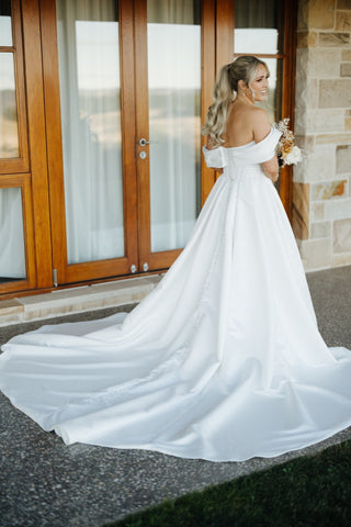 Elegant off-the-shoulder wedding gown with a dramatic satin train, button-back detailing, and timeless bridal style for 2025 weddings.