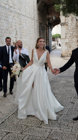 Chic satin wedding dress in ivory with a deep V-neckline, sleeveless design, belted waist, and dramatic floor-length A-line skirt with a leg slit, tailored for contemporary brides