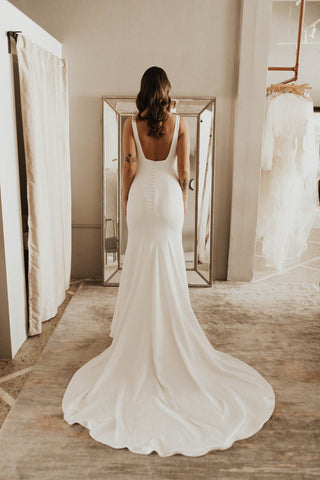 Minimalist crepe wedding gown with a low scoop back, button-down detailing, and a sleek figure-hugging silhouette that flows into a graceful chapel train, perfect for modern and elegant bridal looks