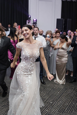 Sophisticated wedding scene showcasing a bride in a floral-appliqué gown with a flowing train and sheer bodice, and a groom in a white tuxedo, walking down an elegant outdoor aisle with scenic mountain views