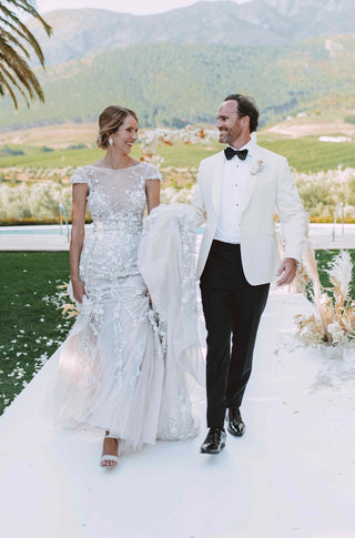 Elegant wedding gown with intricate floral appliqués, sheer illusion neckline, cap sleeves, and flowing train, paired with a groom in a classic white tuxedo and black trousers, set against a stunning outdoor vineyard backdrop