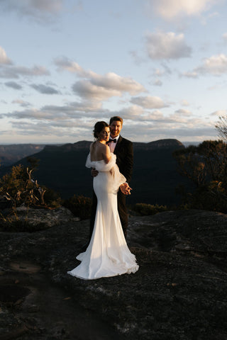 Romantic off-shoulder bridal gown with sheer sleeves, a structured bodice, and a sleek column skirt, ideal for intimate outdoor weddings or timeless bridal styles