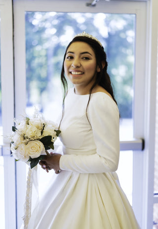 This timeless wedding gown combines classic elegance with modern minimalism. The high scoop neckline and long, graceful sleeves complement the full, flowing skirt, which moves with every step. The dress’s simplicity is its strength, allowing the bride's natural beauty to shine while making a sophisticated statement in any setting, whether indoor or outdoor.