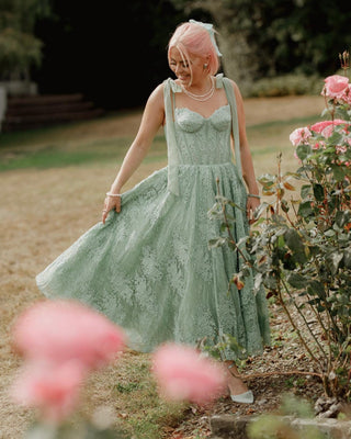 Romantic sage green lace midi dress featuring a corset-style bodice, delicate straps tied in bows, and a flowing A-line skirt, ideal for garden weddings or vintage-inspired events