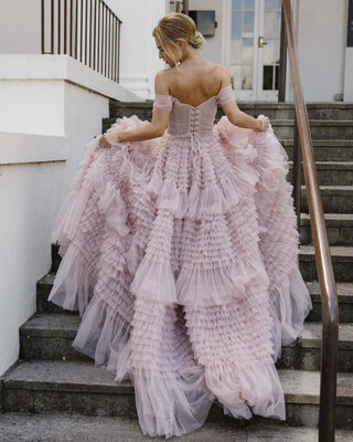 Whimsical blush pink gown featuring off-the-shoulder tulle sleeves, a corset-style lace-up back, and dramatic layered ruffles cascading into a full ballgown silhouette, perfect for fairy-tale weddings or formal event