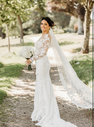 Chic ivory wedding gown with 3D floral lace detailing, deep V-neckline, off-the-shoulder sheer long sleeves, and a dramatic fit-and-flare silhouette, tailored for contemporary bridal aesthetics