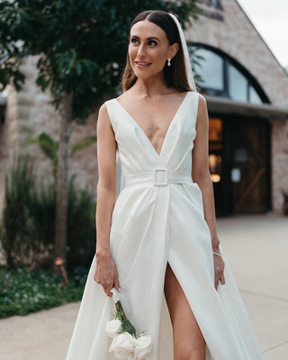 Sophisticated ivory satin wedding dress with a plunging V-neckline, belted waist detail, and flowing A-line silhouette with a slit, designed for stylish and timeless weddings