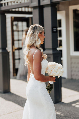 Fitted bridal gown with a shimmering fabric, spaghetti straps, and a graceful train, offering timeless elegance with a modern edge