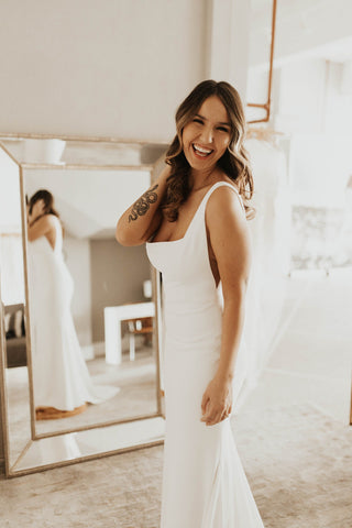 Chic and timeless bridal dress featuring a clean, minimalist design, low back, and delicate row of buttons, complemented by a soft, flowing train, ideal for understated yet sophisticated weddings