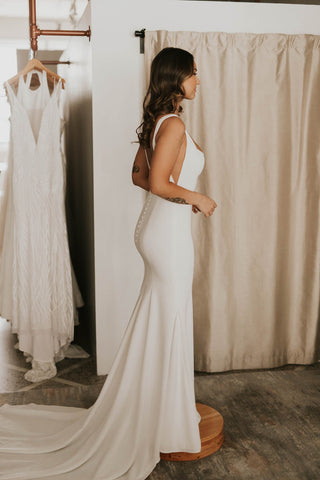 Elegant white wedding gown with a flattering fitted silhouette, scoop back with button accents, and a fluid train, perfect for contemporary brides seeking simplicity and style