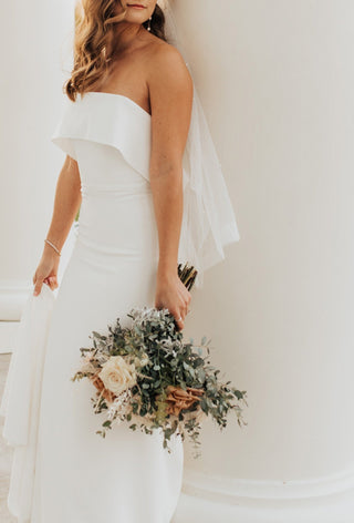 Modern strapless wedding dress with a sleek silhouette, minimalistic design, featuring a chic ruffled neckline, perfect for contemporary brides seeking elegance, paired with a delicate pearl veil and romantic bouquet